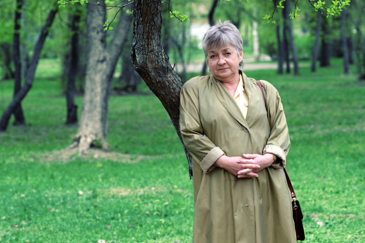 kobieta w wieku 60 plus w płaszczu stoi na zewnątrz na tle zieleni z poważną miną