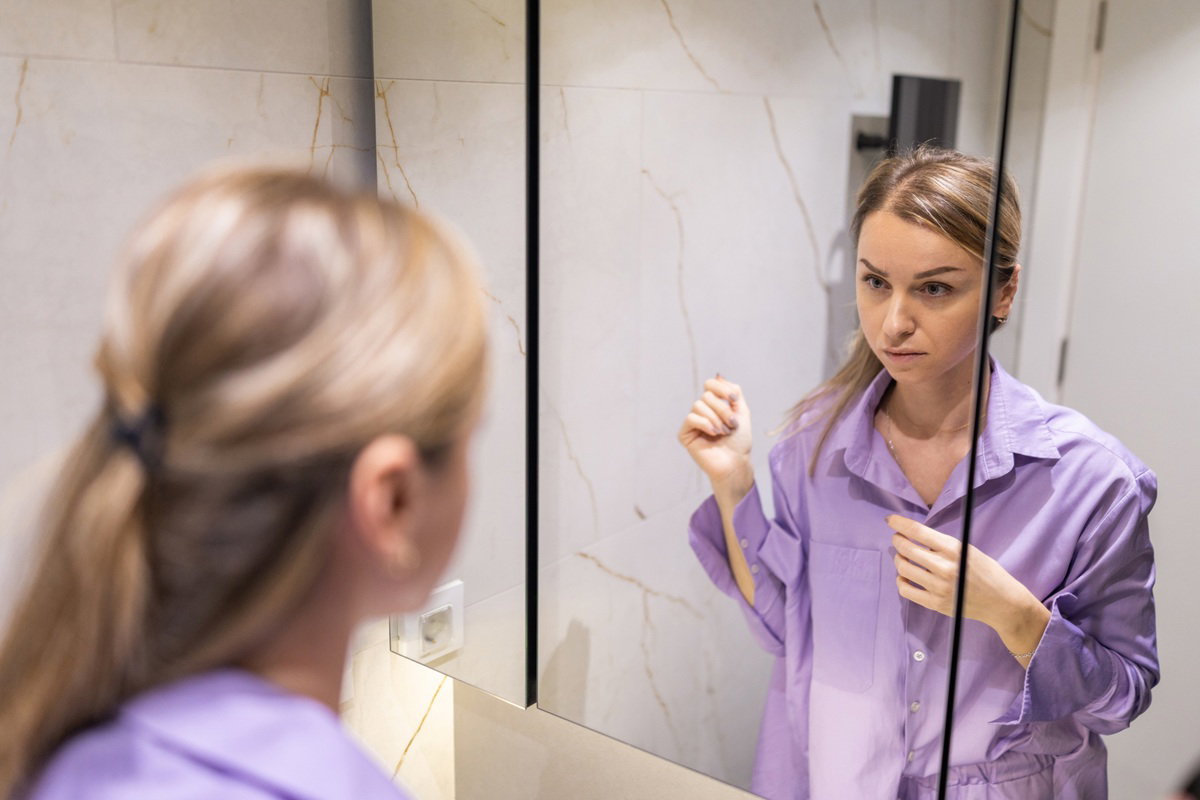 kobieta w wieku 30 plus w fioletowej koszuli patrzy na siebie w lustrze z poważną miną