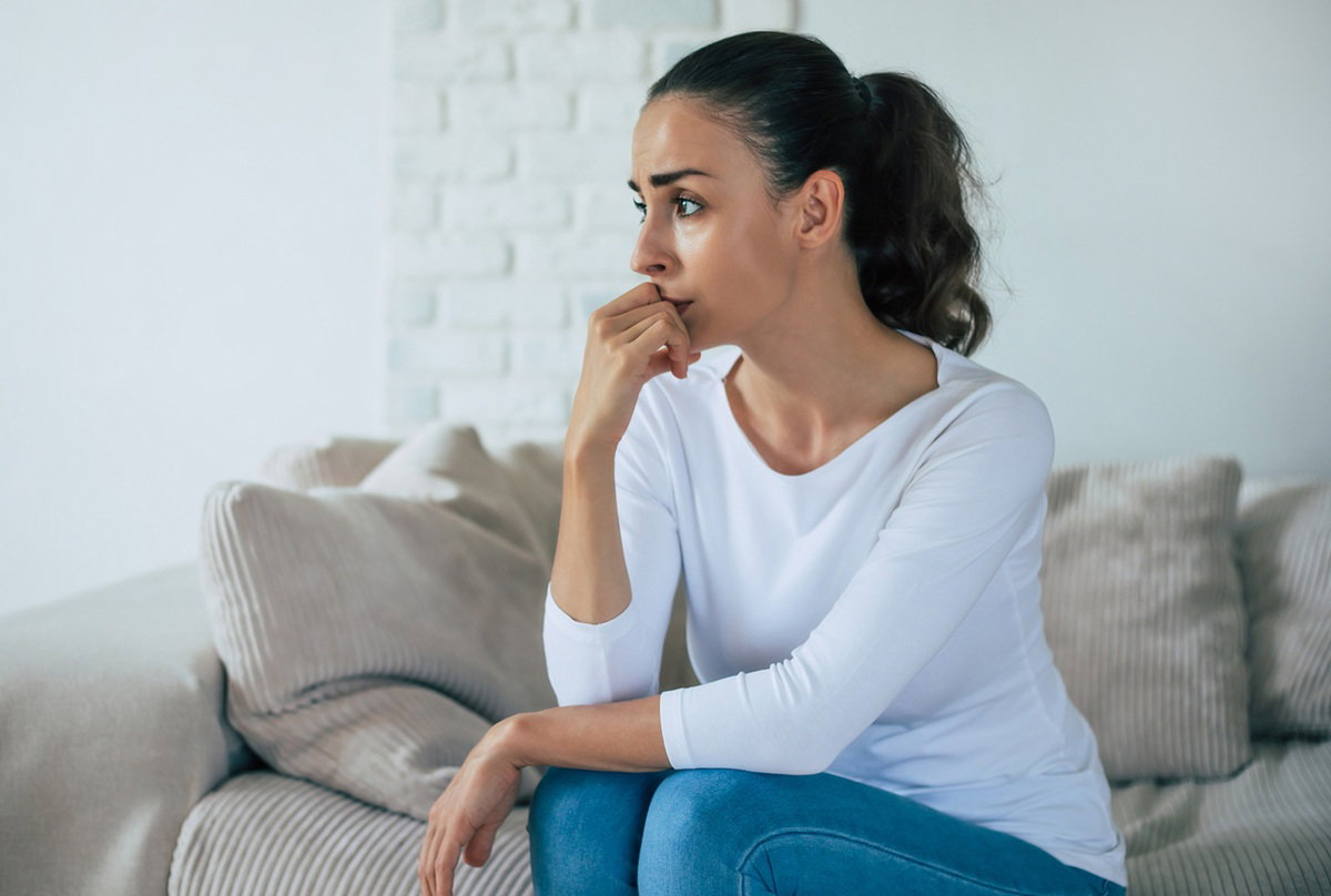 kobieta w wieku 30 plus ciemne włosy w białej bluzce siedzi na kanapie zmartwiona