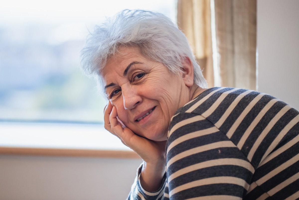 kobieta w wieku 65 plus w bluzce w paski siwa krótko ostrzyżona siedzi przy oknie
