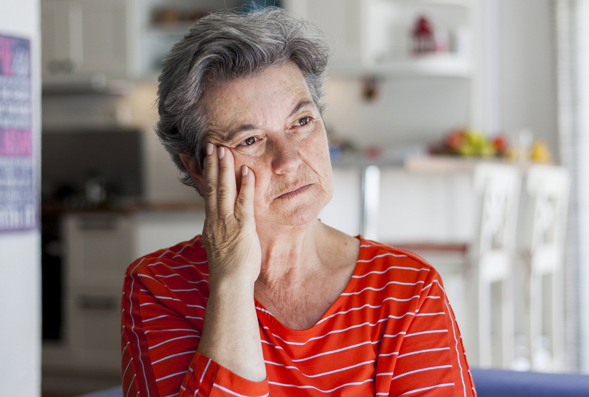 kobieta w wieku 60 lat w czerwonej bluzce w prążki siedzi zmartwiona na tle kuchni