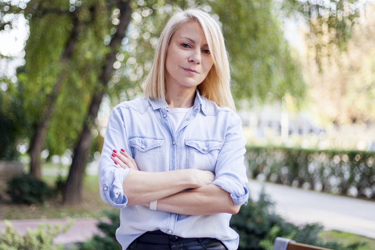 kobieta w wieku 30 plus blondynka stoi na tle drzew z lekko kpiącym gorzkim uśmiechem