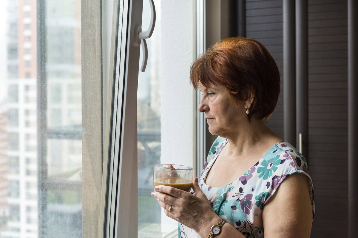 kobieta w wieku 55 plus stoi przy oknie z kawą zamyślona i patrzy w dal