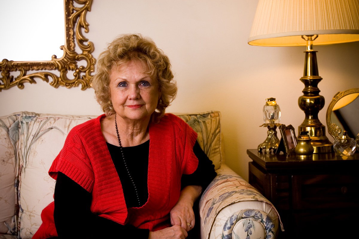 kobieta w wieku 60 plus w czerwonym blezerze siedzi na kanapie z lekkim uśmieszkiem