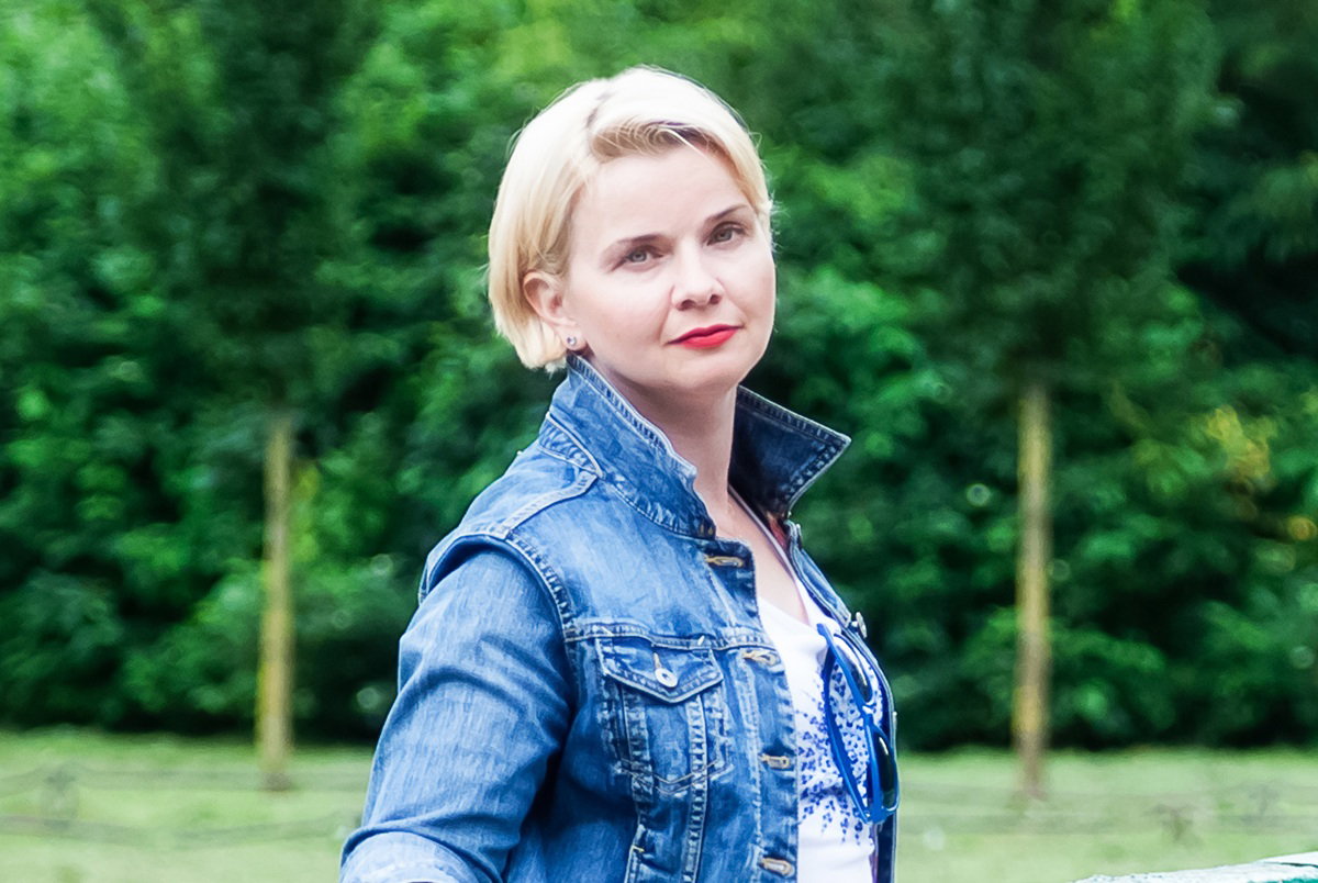kobieta w wieku 40 plus w jeansowej kurtce stoi na tle zieleni zamyślona