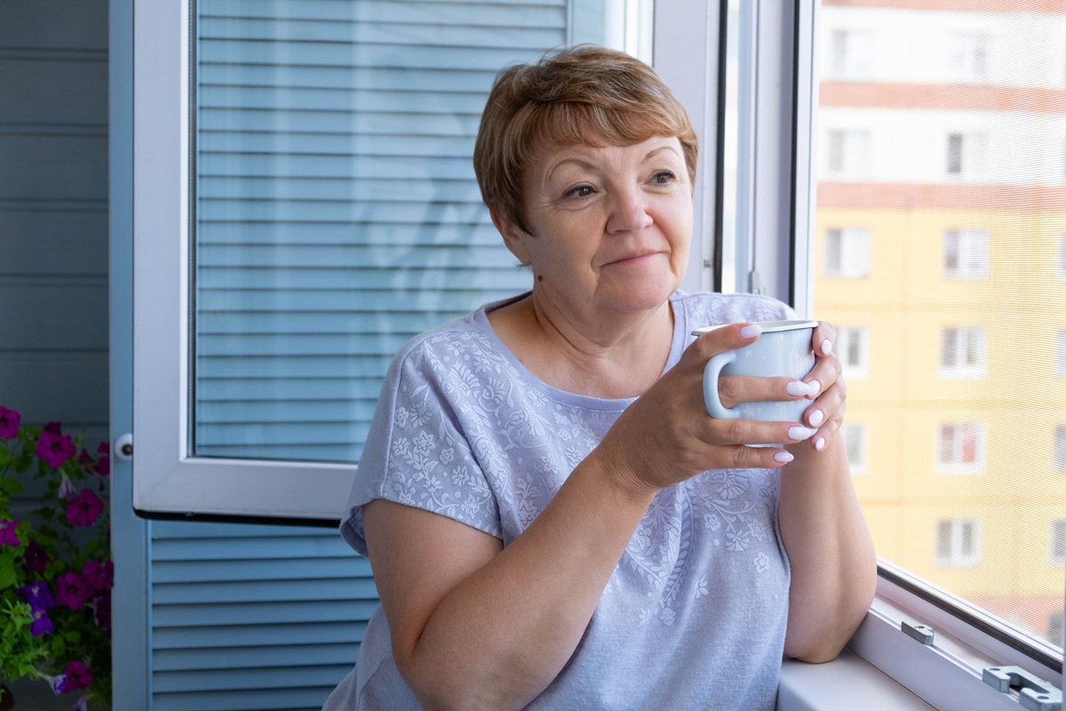 kobieta w wieku 60 plus z krótkimi włosami stoi na balkonie z kubkiem herbaty zamyślona
