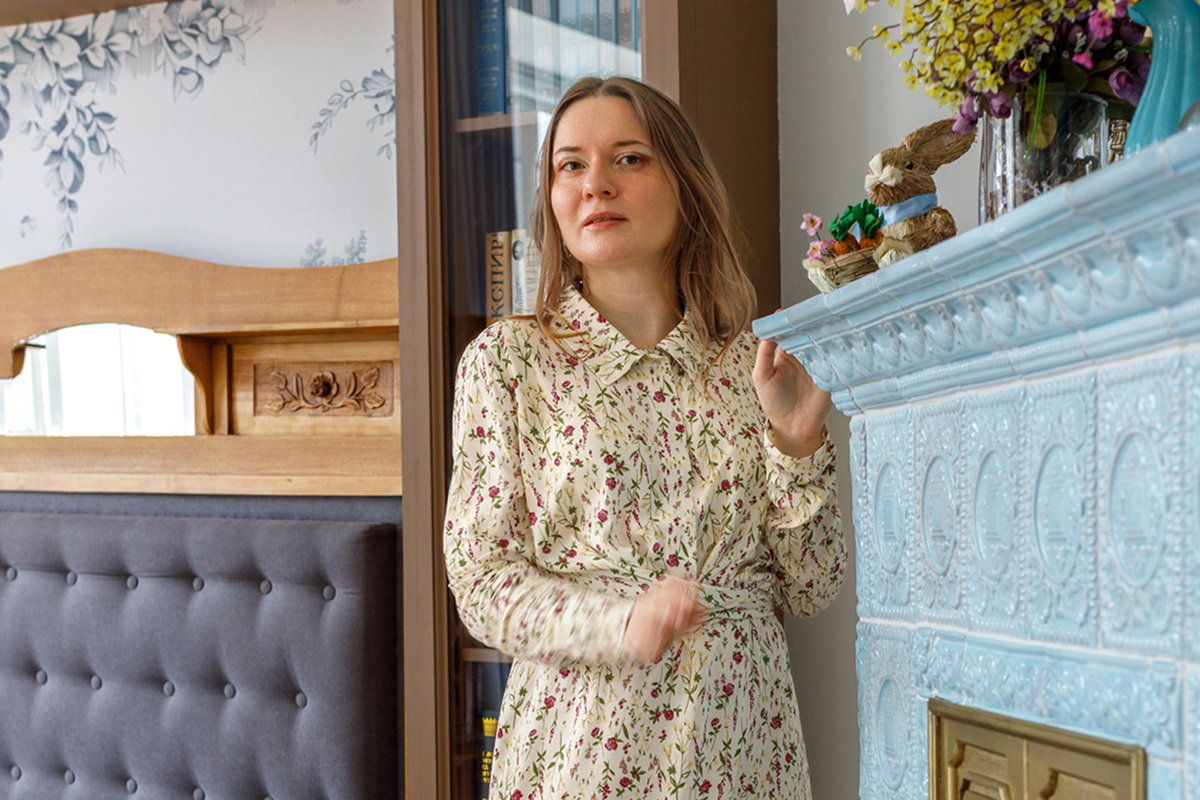 kobieta w wieku 30 plus stoi przy kaflowym błękitnym piecu zamyślona w kwiecistej sukience