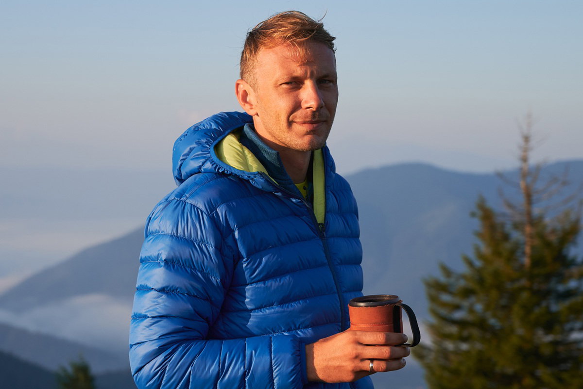 mężczyzna w wieku około 35 plus w niebieskiej kurtce stoi na tle gór z kubkiem w ręku zamyślony