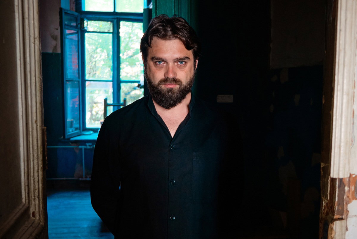 mężczyzna w wieku 30 plus z wąsami i brodą stoi w starym domu z poważną miną