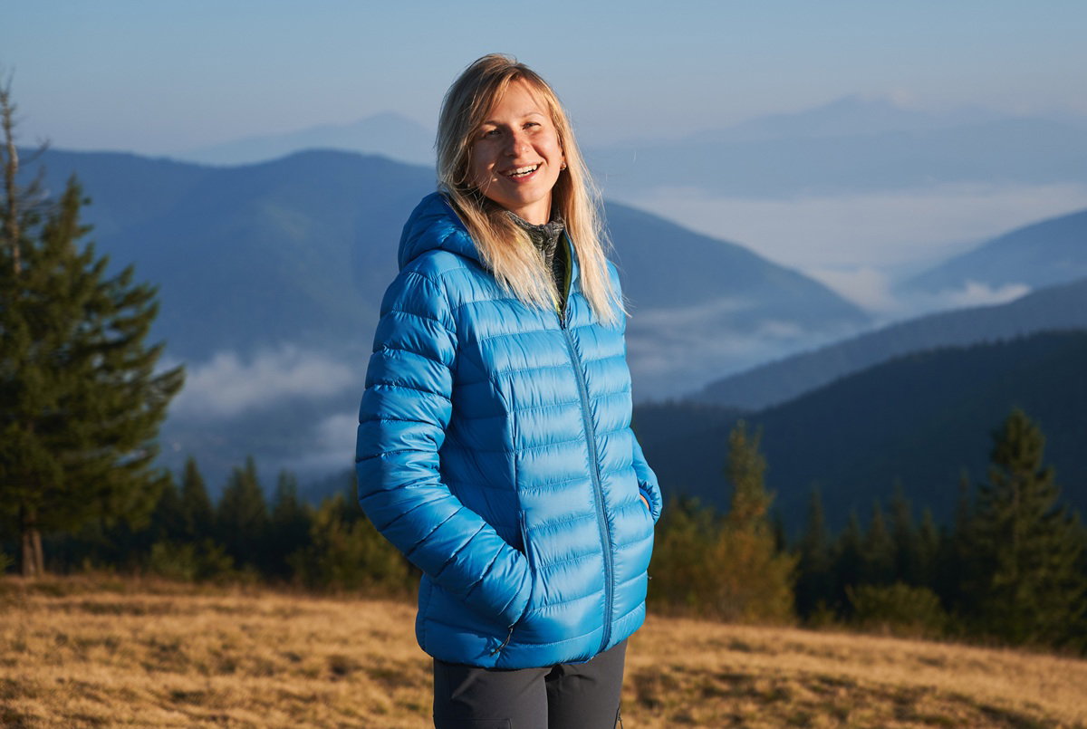 kobieta w wieku około 30 lat w niebieskiej kurtce stoi na tle gór i się uśmiecha