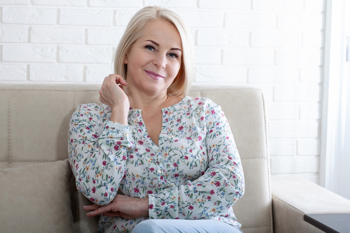 kobieta w wieku 40 plus blondynka w kwiecistej bluzce siedzi na kanapie z lekkim uśmiechem