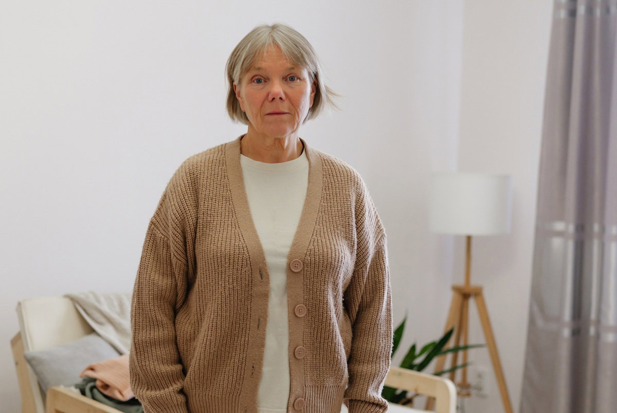 kobieta w wieku 55 plus z siwymi włosami w beżowym swetrze stoi smutna na tle salonu