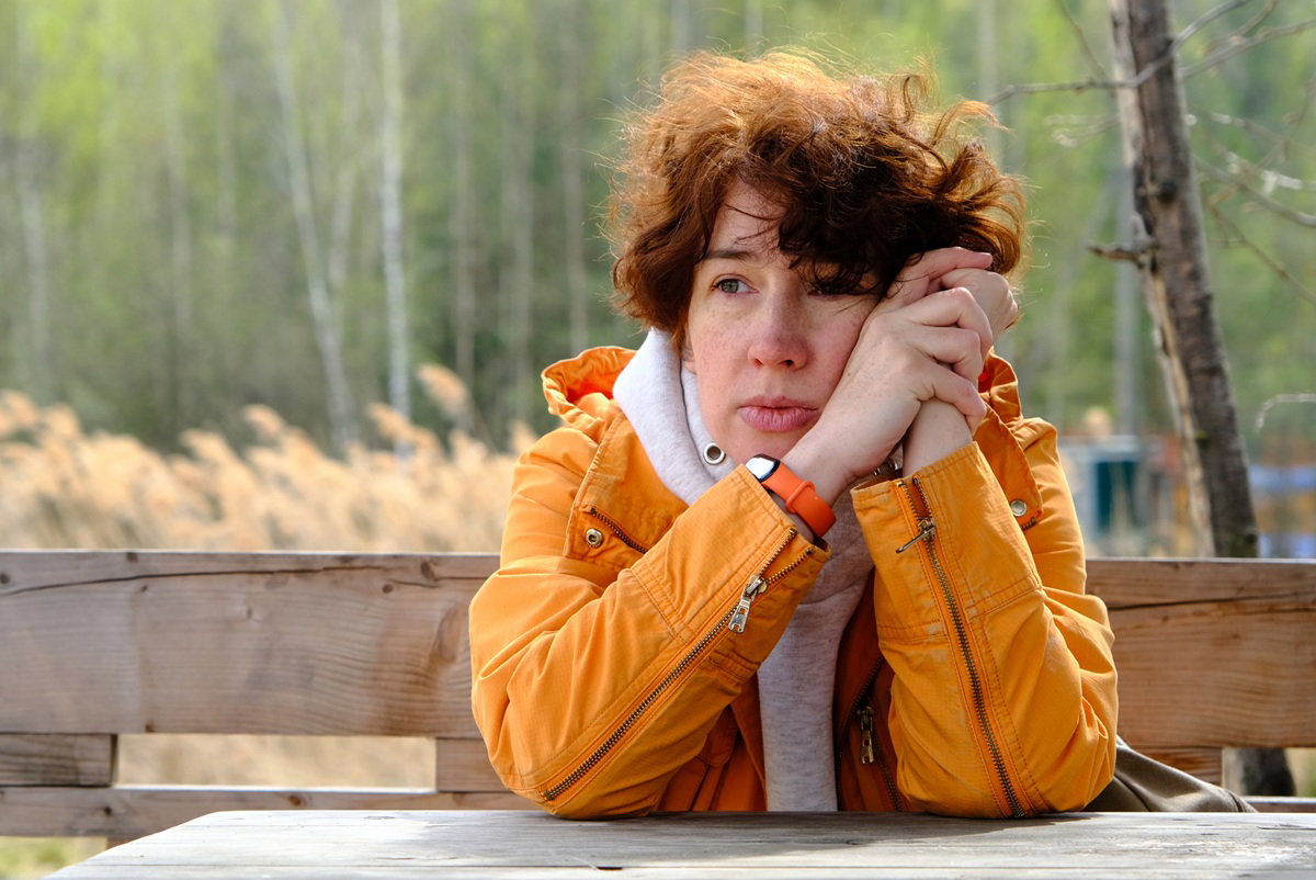 kobieta w żółtej kurtce w wieku 30 plus siedzi przy drewnianym stole zamyślona na tle lasu