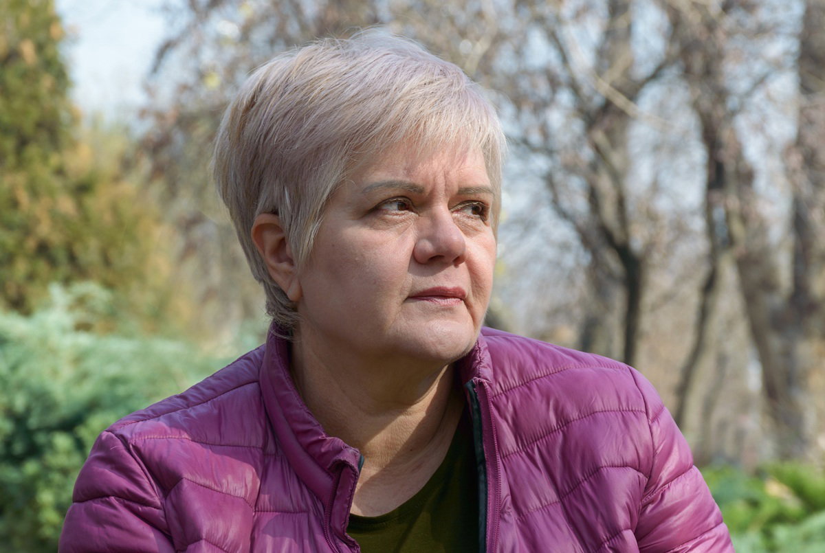 kobieta w wieku 55 plus z krótką fryzurą i w fioletowej kurtce siedzi na zewnątrz i podejrzliwie patrzy