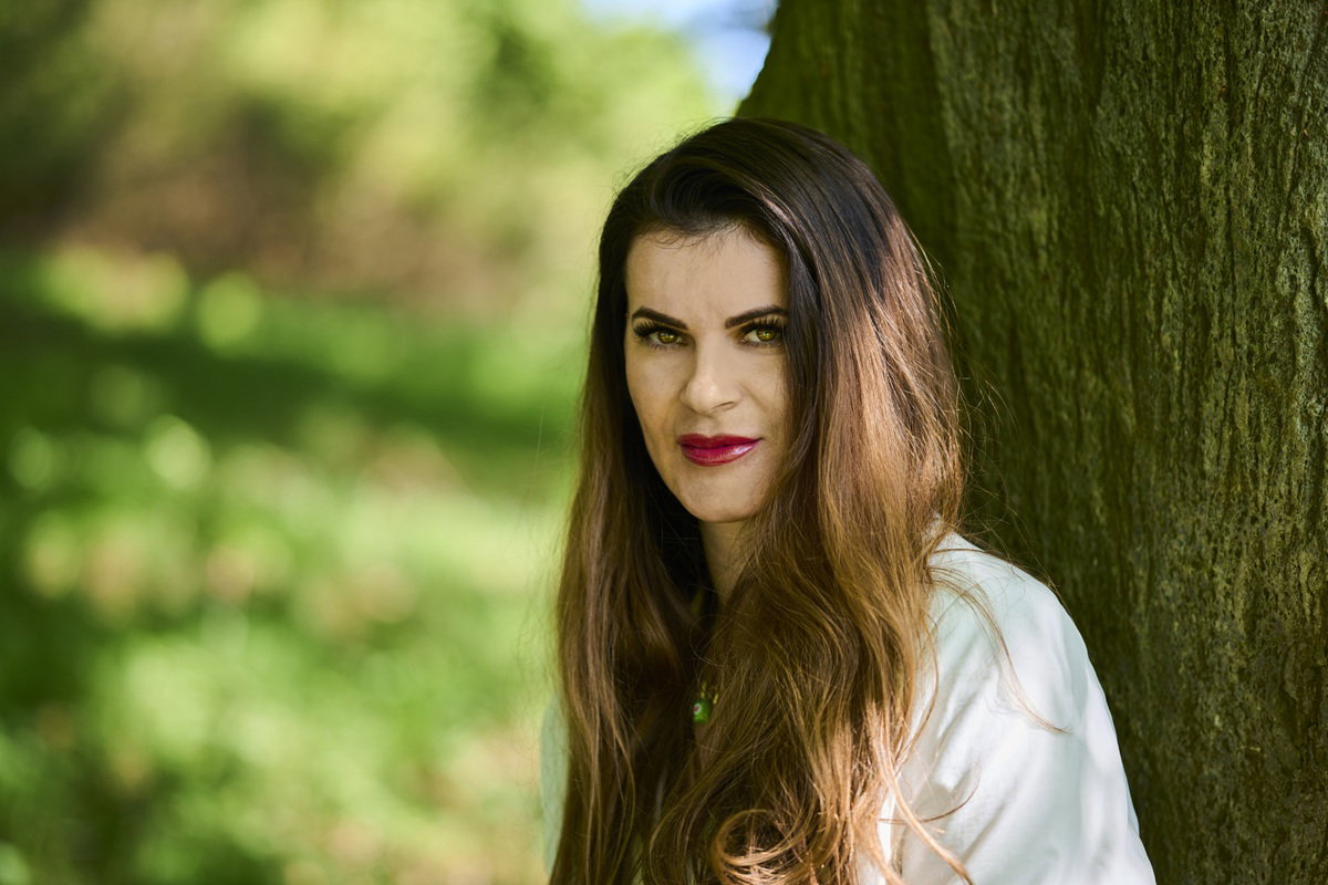 kobieta w wieku 30 plus w makijażu stoi na tle drzewa z lekkim uśmieszkiem