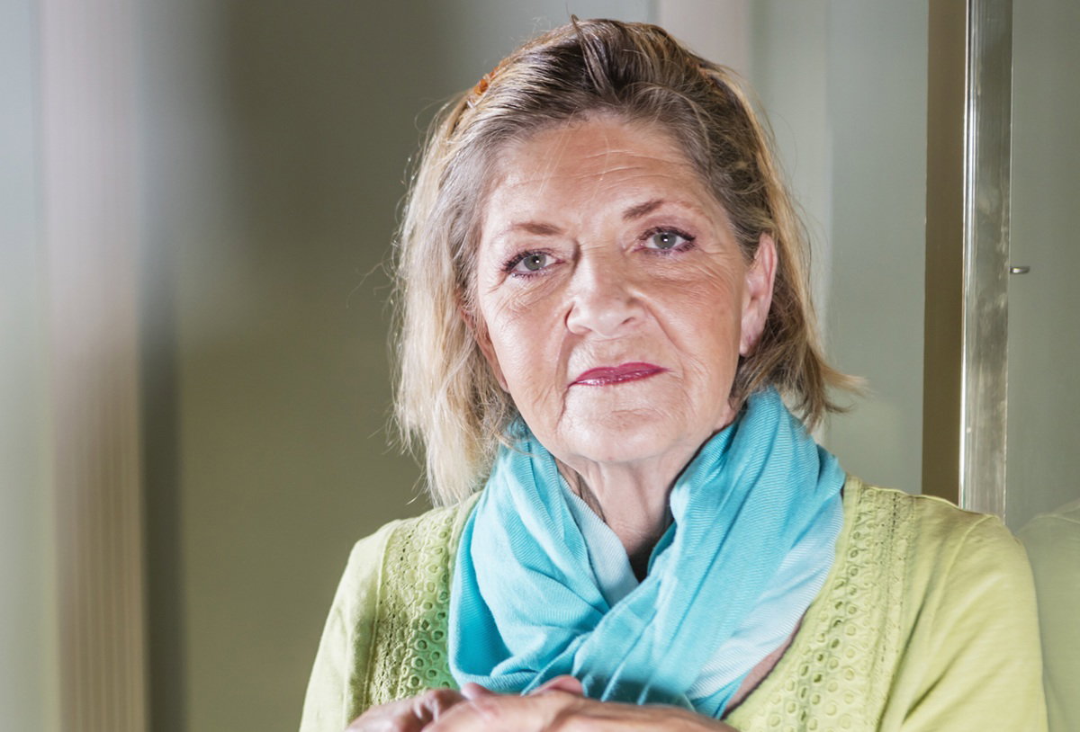 kobieta w wieku 60 plus w niebieskiej apaszce na szyi stoi wewnątrz z poważną miną