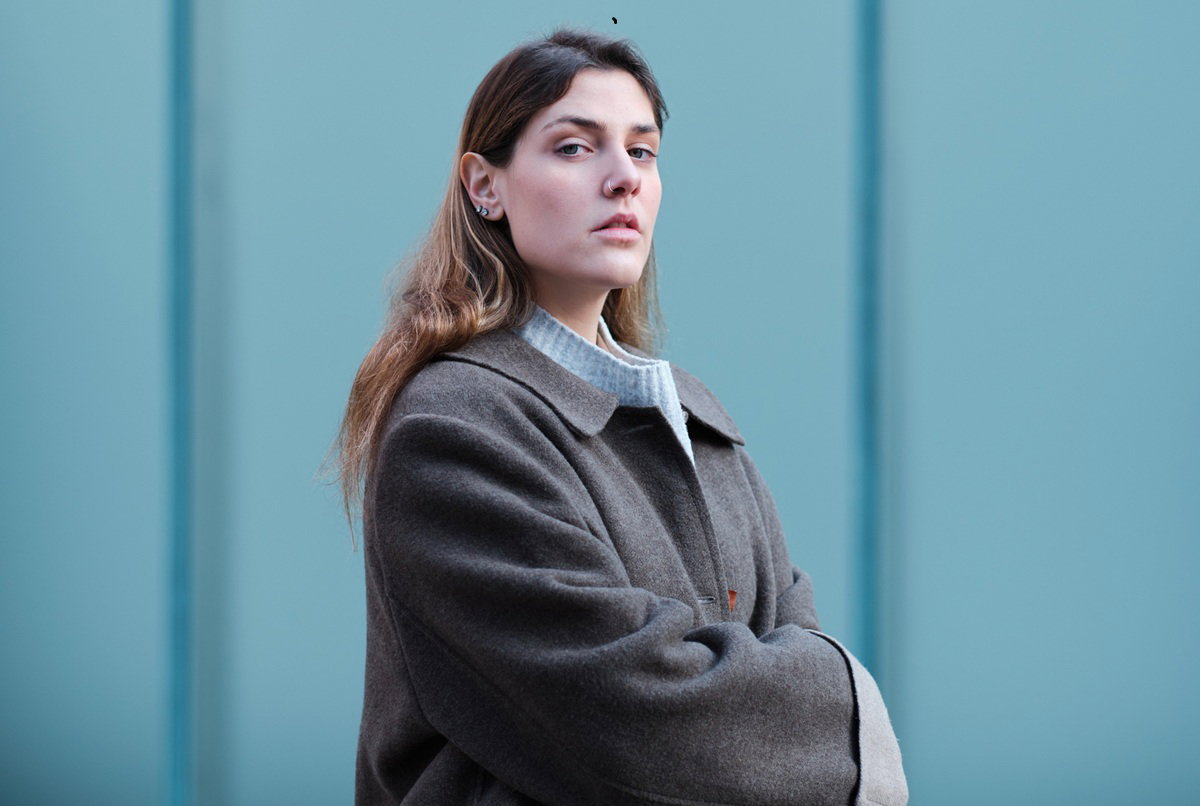 kobieta w wieku 30 plus w szarym płaszczu patrzy dumnie przed siebie na tle niebieskiej ściany