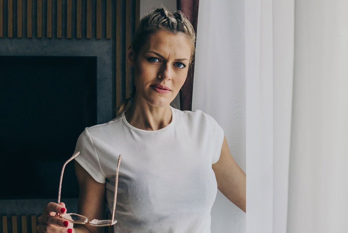 kobieta w wieku 40 plus blondynka stoi przy oknie z okularami w ręku ze zmieszaną miną