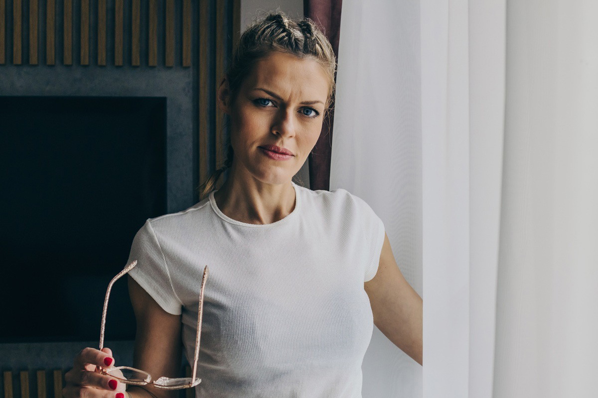 kobieta w wieku 40 plus blondynka stoi przy oknie z okularami w ręku ze zmieszaną miną