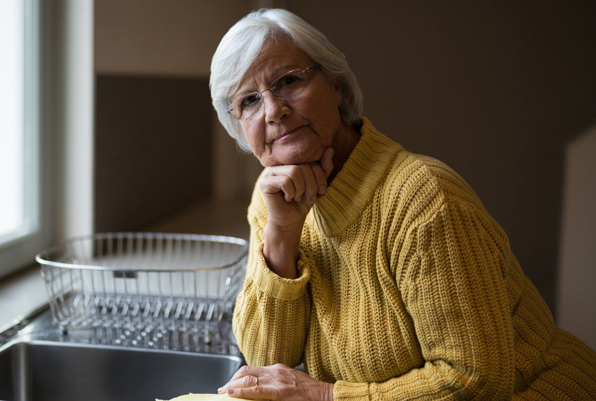 kobieta w wieku 60 plus w żółtym swetrze siwe włosy i okulary stoi nad zlewem zmartwiona