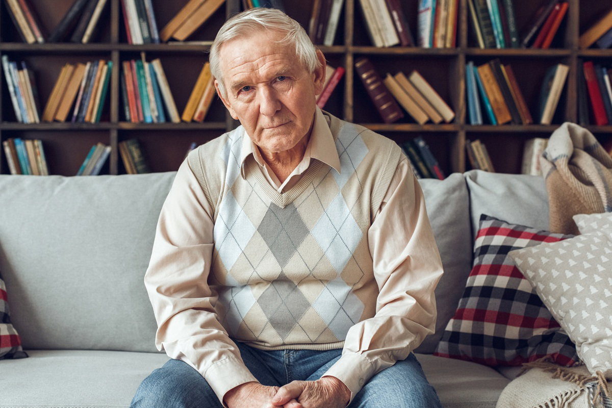 mężczyzna w wieku 65 plus siwy w pulowerze w klasyczny wzór siedzi na kanapie zdruzgotany