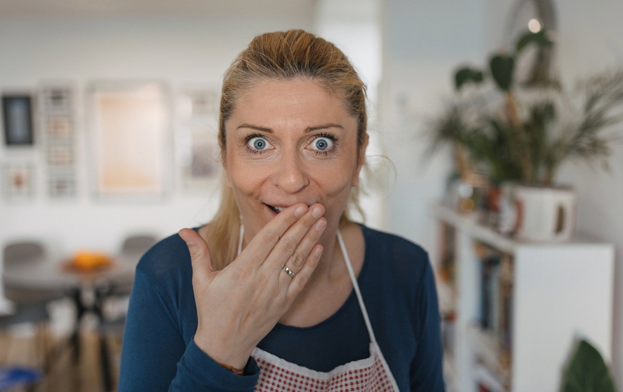 kobieta w wieku 40 plus w fartuszku stoi na tle jadalni i śmieje się skrycie z wesołymi oczami