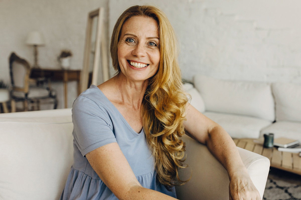 kobieta w wieku 45 plus w niebieskiej sukience siedzi w salonie i szeroko się uśmiecha