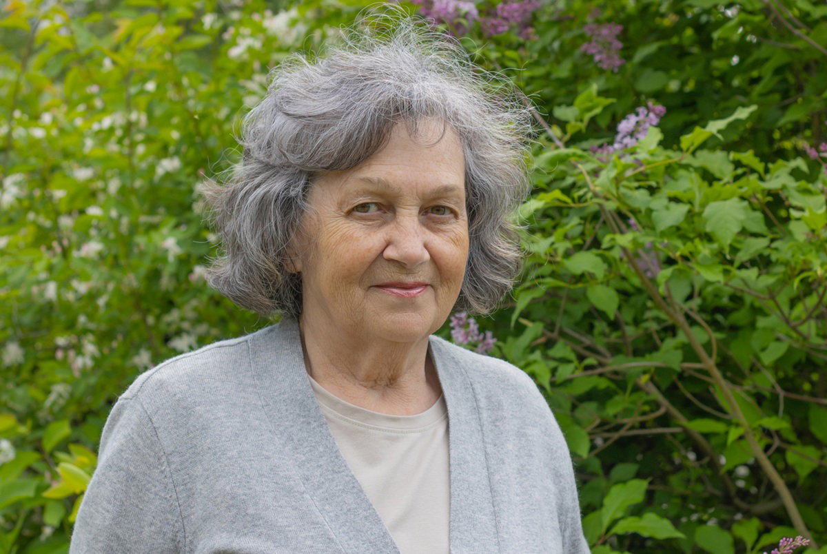 kobieta w wieku 65 plus siwe włosy w szarym swetrze stoi na tle bujnych roślin