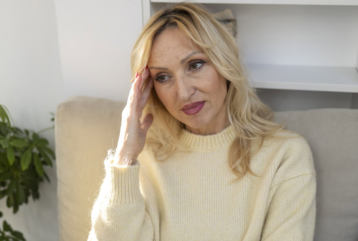 kobieta w wieku 50 plus blondynka w jasnym swetrze siedzi zmartwiona na sofie