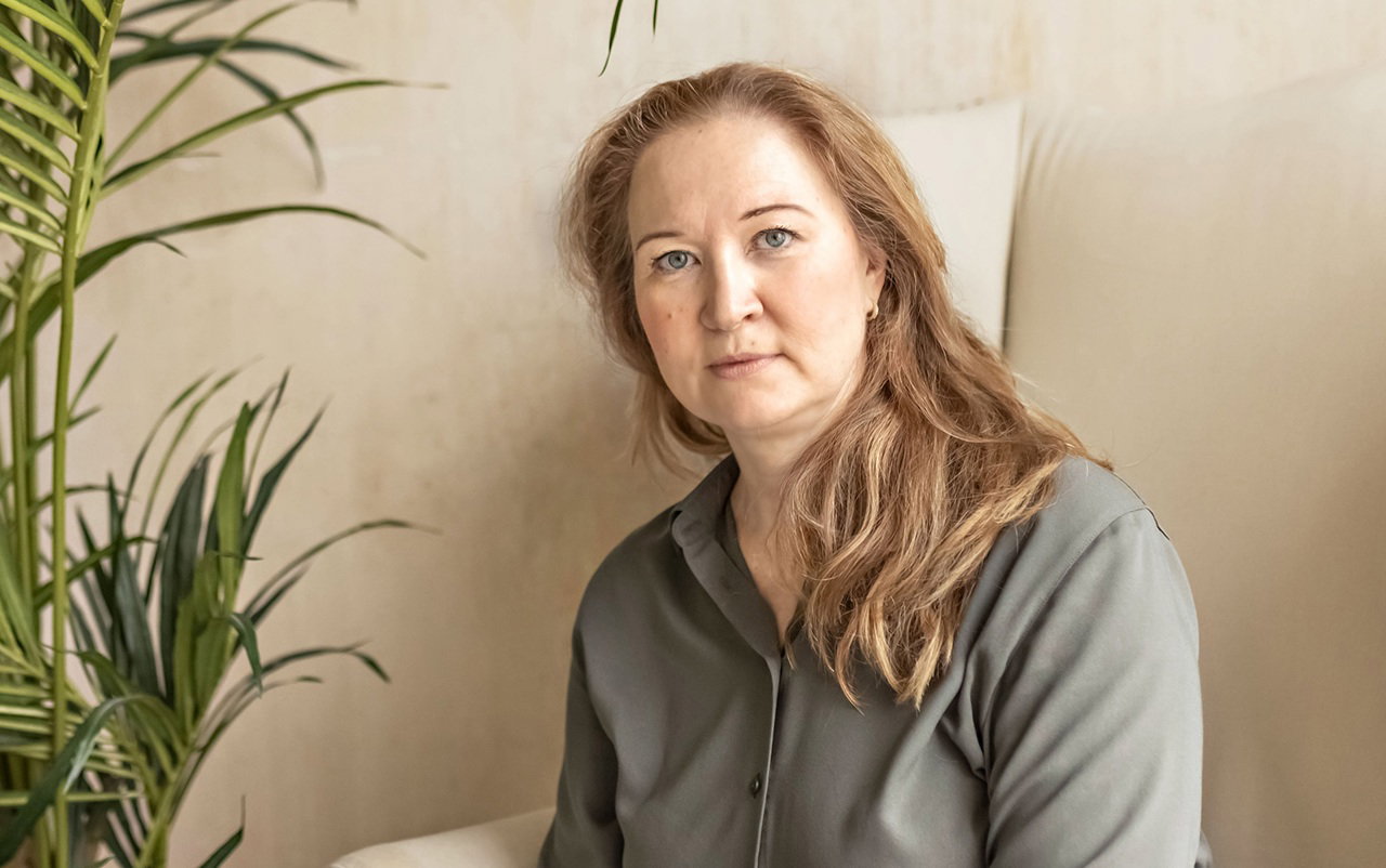 kobieta w wieku 40 plus w szarej koszuli siedzi na jasnym fotelu z poważną miną