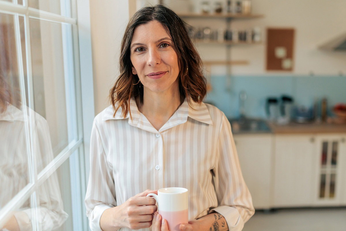 kobieta w wieku 40 plus w koszuli w paski stoi na tle kuchni z kubkiem w ręku i lekkim uśmieszkiem