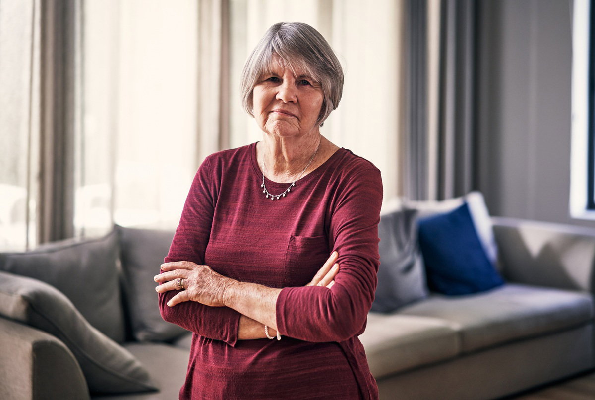 kobieta w wieku 70 plus w ciemnoczerwonej bluzce stoi na tle mieszkania urządzonego w szarościach