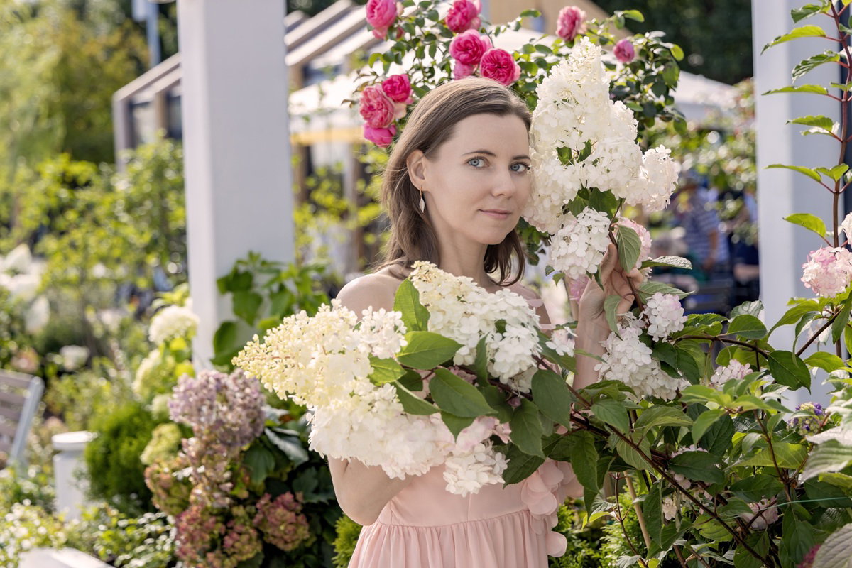 kobieta w wieku 35 plus w ogrodzie wśród kwiatów hortensji i róż stoi z poważną miną