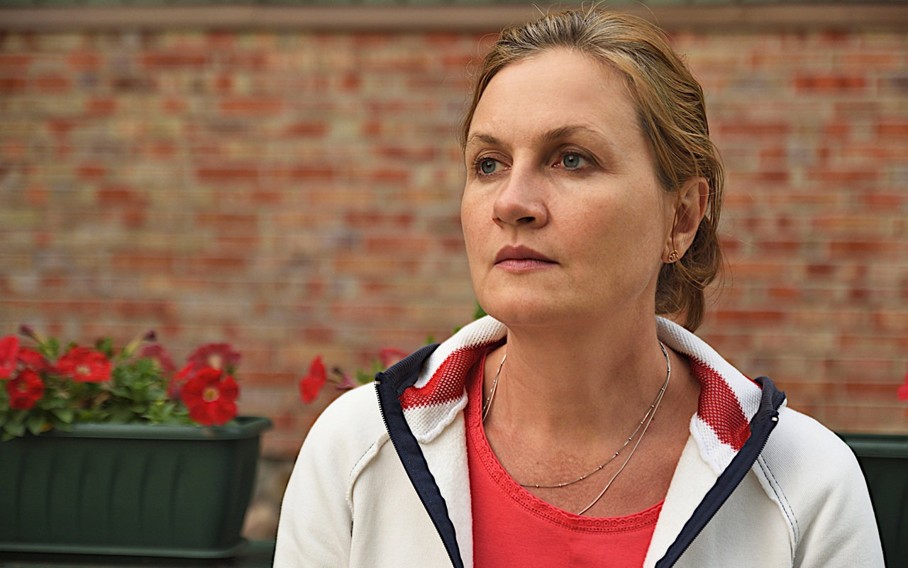 kobieta w wieku 40 plus w jasnej bluzie siedzi na tle ściany w ogrodzie smutna