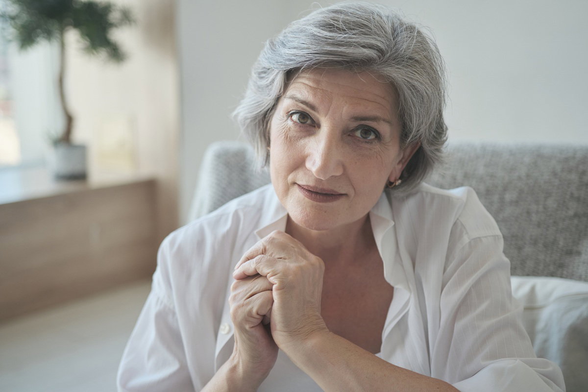 kobieta w wieku 55 plus z siwymi włosami w białej koszuli z kpiącą miną patrzy przed siebie