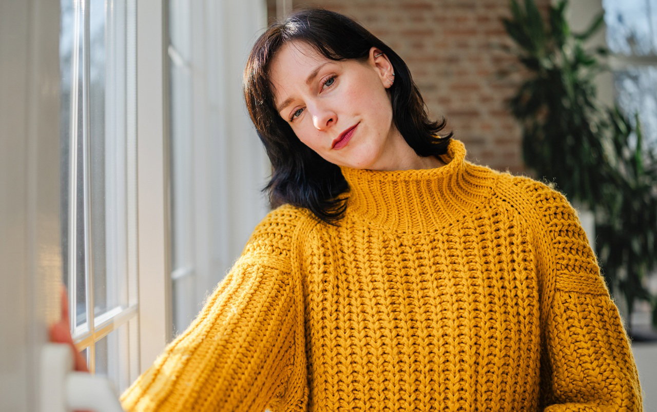 kobieta w wieku 35 plus w żółtym swetrze stoi przy drzwiach balkonowych ze smutną miną