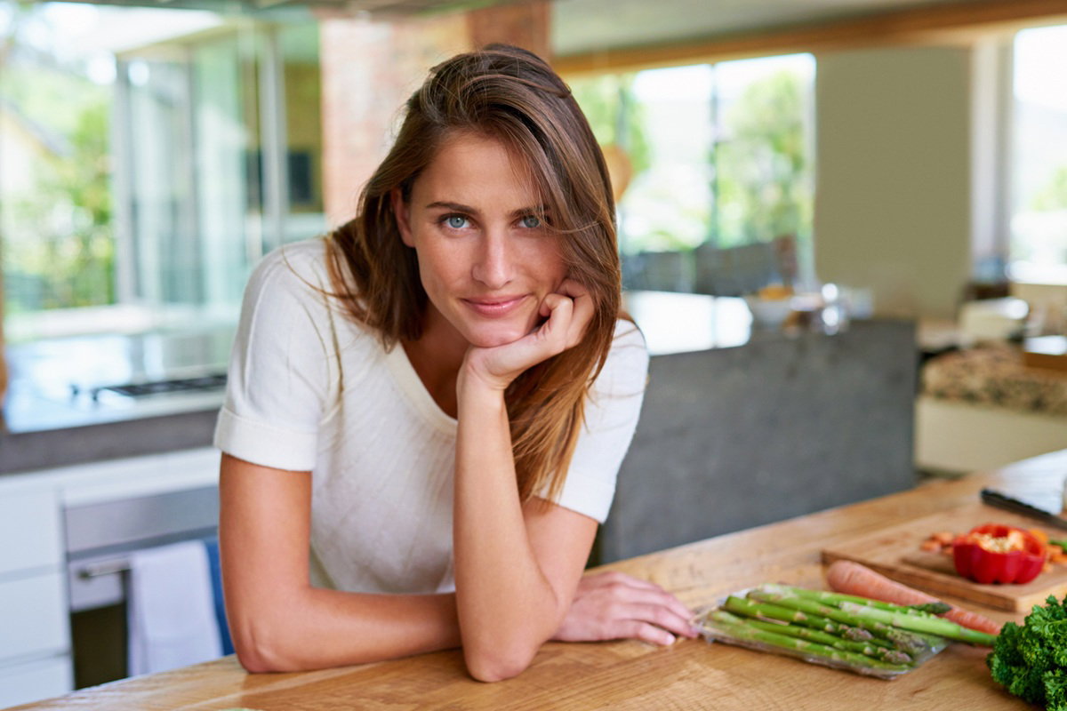 kobieta w wieku 30 plus stoi w kuchni z zadziornym spojrzeniem a na blacie leżą szparagi