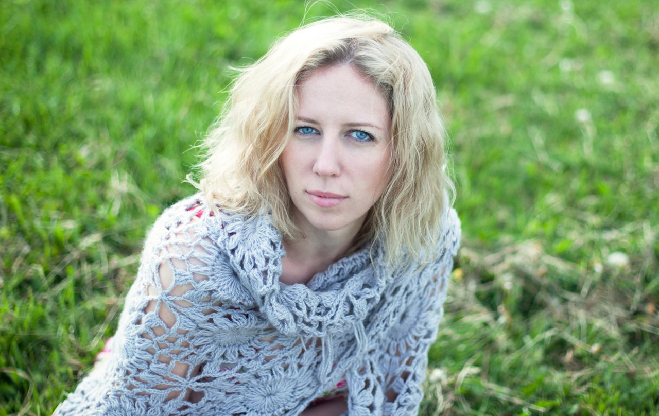 kobieta w wieku 35 plus blondynka siedzi na trawie w szarej dzierganej chuście z poważną miną