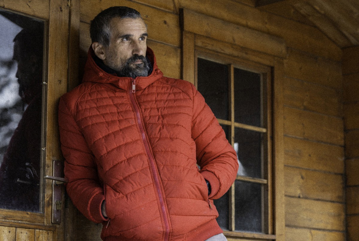mężczyzna w wieku 45 plus w czerwonej kurtce z brodą i wąsami stoi na tle drewnianej chaty zasępiony