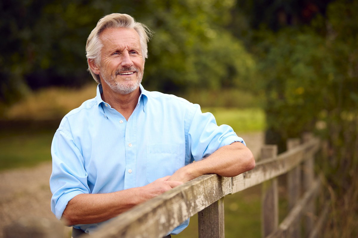 mężczyzna w wieku 65 lat w niebieskiej koszuli stoi przy płocie na tle zieleni uśmiechnięty