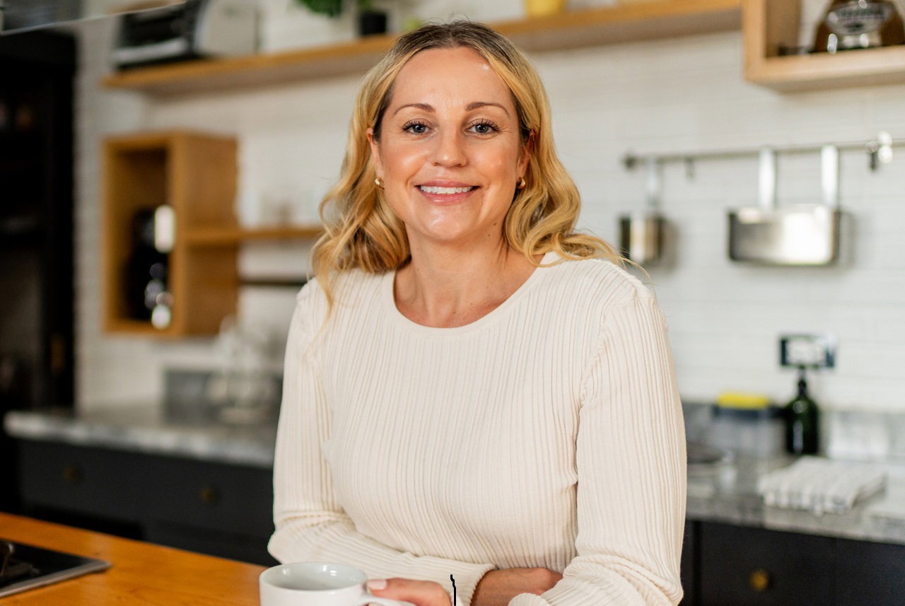 kobieta w wieku 30 plus blondynka stoi na tle kuchni z kubkiem w ręku i się uśmiecha kpiąco