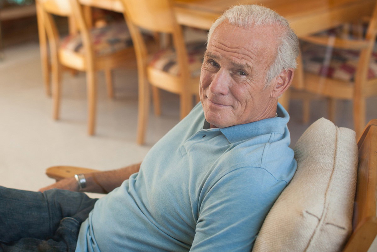 mężczyzna w wieku 65 plus siwiejący i łysiejący siedzi na fotelu na tarasie i uśmiecha się z przekąsem