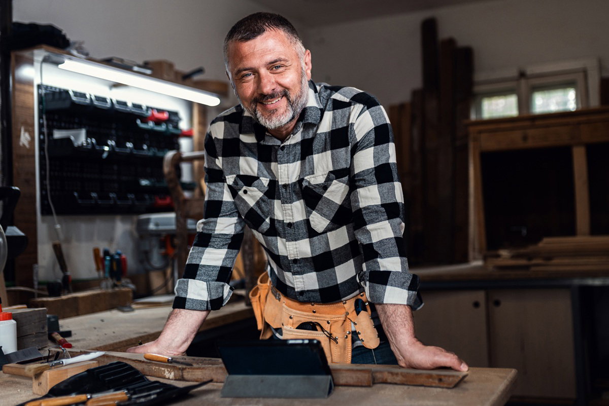 mężczyzna w wieku 40 plus w kraciastej koszuli stoi w warsztacie i się uśmiecha