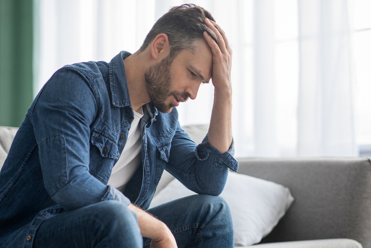 mężczyzna w wieku 45 plus z wąsami i brodą w jeansowej koszuli siedzi na kanapie załamany