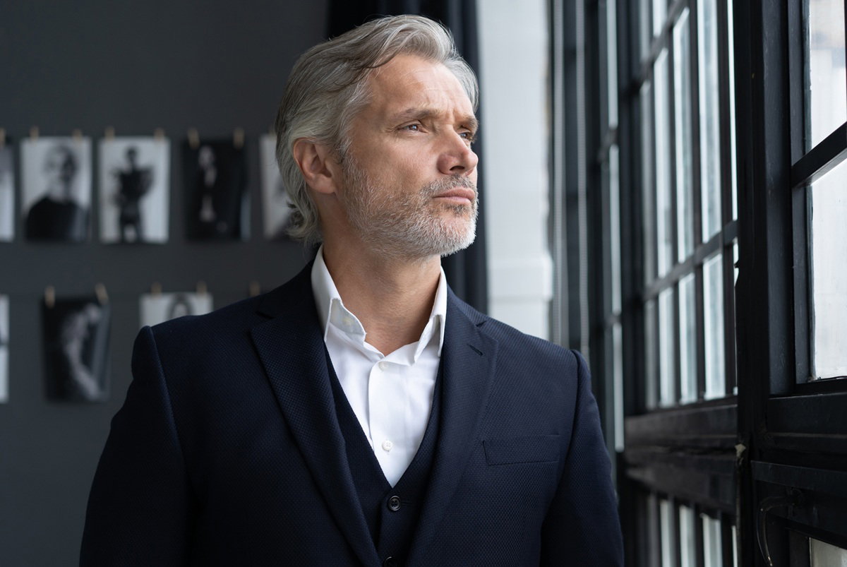 mężczyzna w wieku 40 plus siwy z zarostem w garniturze patrzy zamyślony przez okno