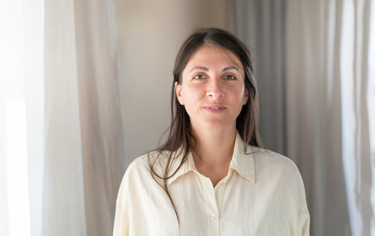 kobieta w wieku 35 plus w jasnej koszuli stoi na tle zasłon obok balkonu z lekkim uśmiechem