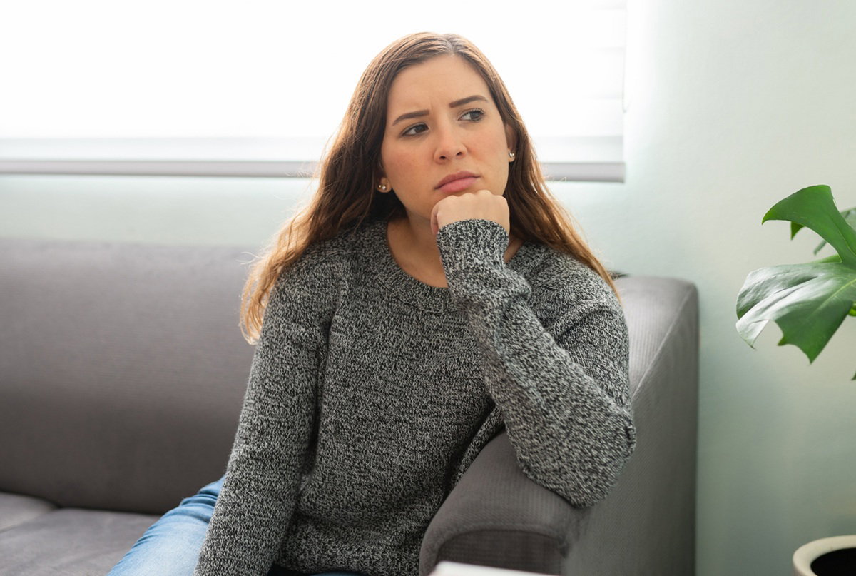 kobieta w wieku 25 plus w szarym melanżowym swetrze siedzi na sofie zmartwiona i zamyślona