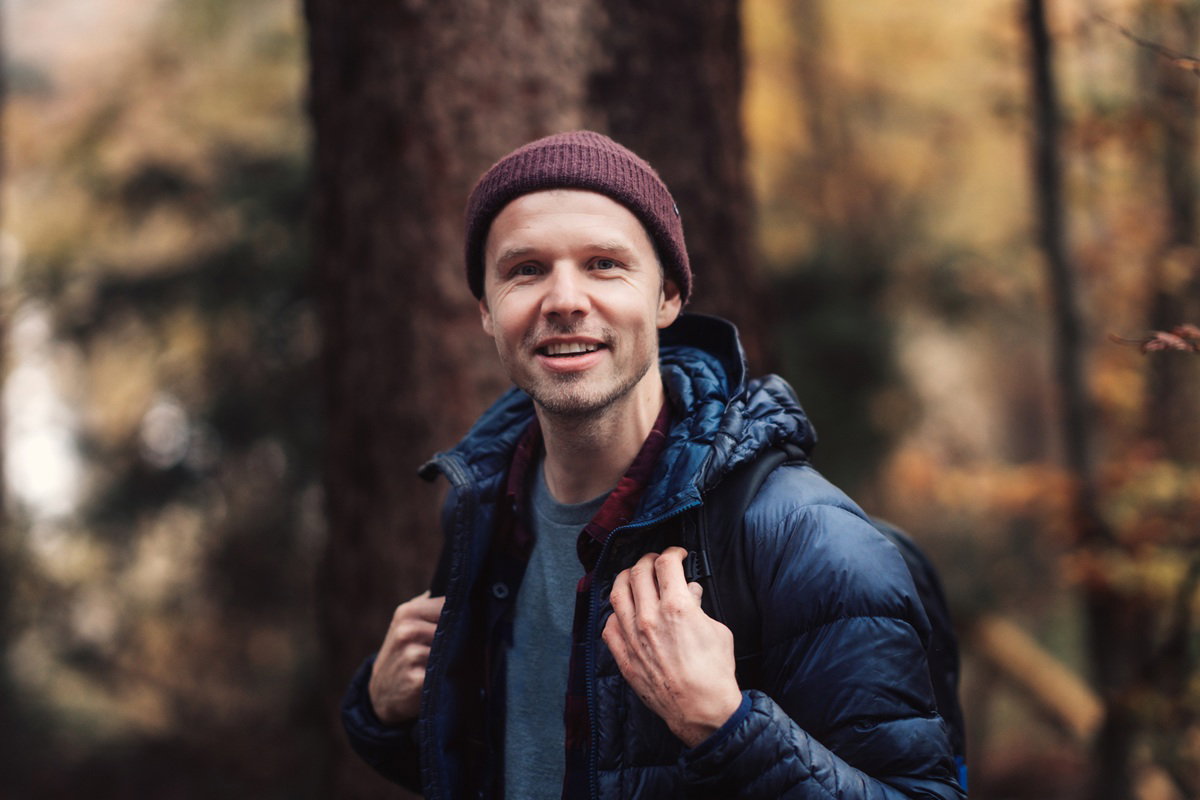 mężczyzna w wieku 35 plus w grantowej kurtce i bordowej czapce stoi w lesie uśmiechnięty