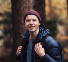 mężczyzna w wieku 35 plus w grantowej kurtce i bordowej czapce stoi w lesie uśmiechnięty