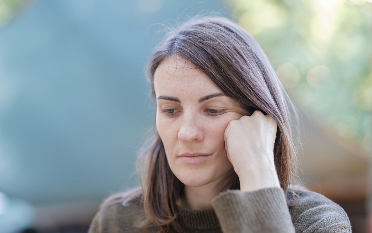kobieta w wieku 35 plus w szaro-burym swetrze siedzi zamyślona i smutna na zewnątrz